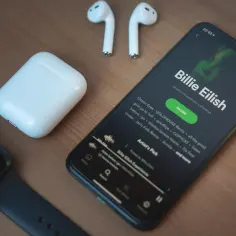 iPhone z aplikacją Spotify, słuchawki AirPods i smartwatch na drewnianym tle.
