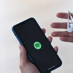 Dłoń trzyma smartfon z logo Spotify, a w drugiej dłoni słuchawki.